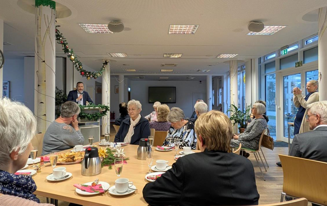 Foto: mehrere Personen an Tischen und ein Redner am Pult