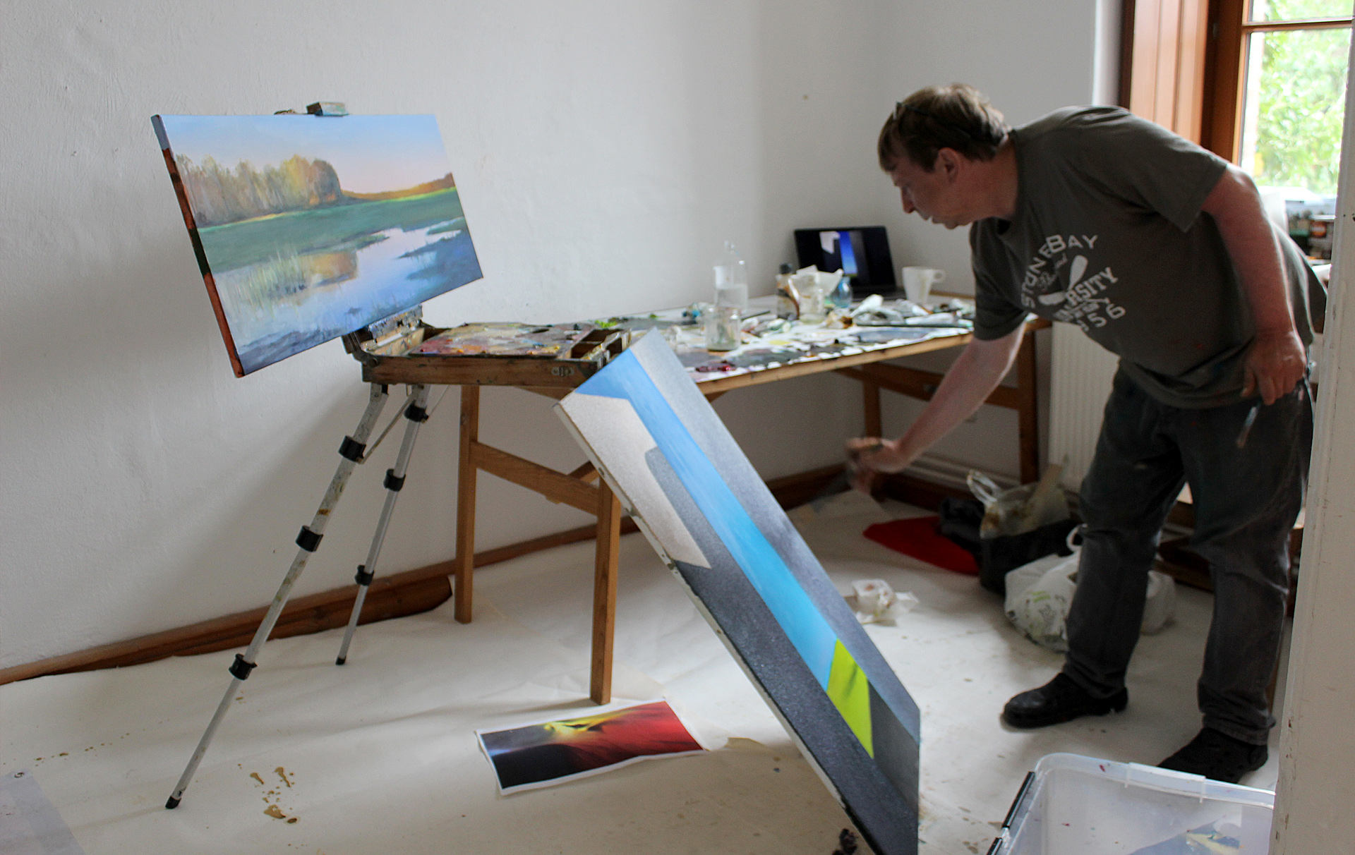 Foto: Künster arbeitet an einem abstrakten Bild, das auf dem Boden steht. Ein 2. Bild mit einer Landschaft steht auf einer Staffelei dahinter Foto: Künster arbeitet an einem abstrakten Bild, das auf dem Boden steht. Ein 2. Bild mit einer Landschaft steht auf einer Staffelei dahinter