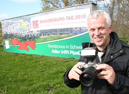 Foto: Polzehl mit Fotoapparat vor der Mauer mit der BRANDENBURG-TAG-Werbung