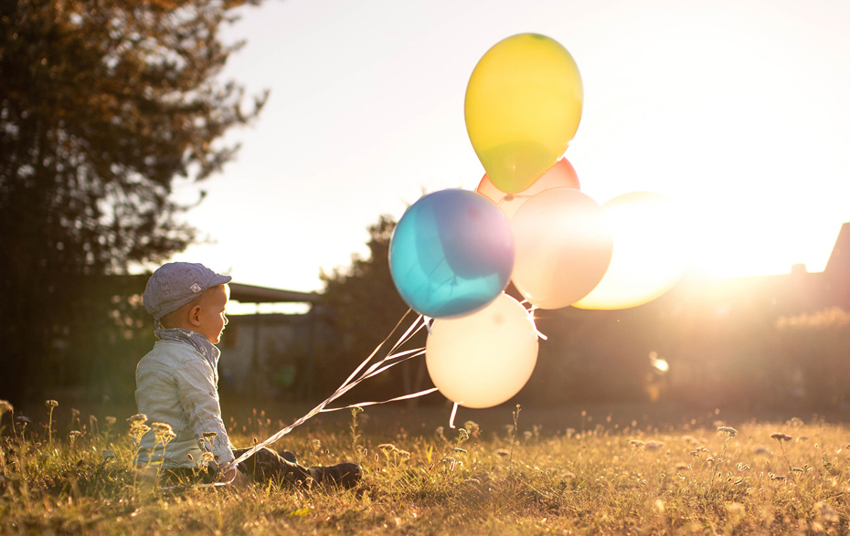 Foto: sitzendes Kind mit mehreren Luftballons im Gegenlicht der Sonne