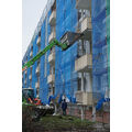 Foto: Eingerüsteter Wohnblock an dem Bauarbeiter mit Bagger Abrissarbeiten vornehmen.