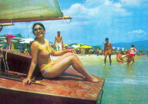 Die Postkarte zeigt eine Frau, auf einem Boot sitzend, am gut gefüllten Badestrand.